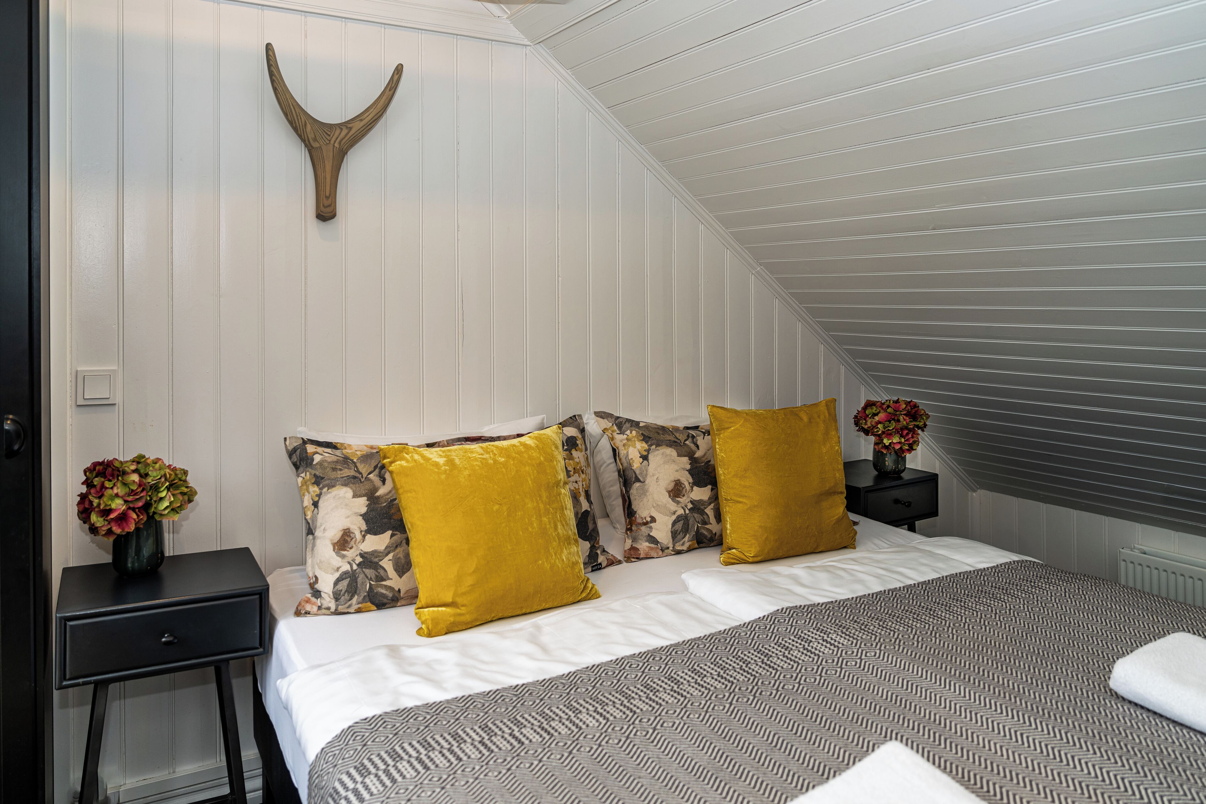 Quarto com teto inclinado, cama com roupa de cama estampada, duas mesas de cabeceira com almofadas florais e amarelas, e um objeto decorativo de madeira fixado na parede.
