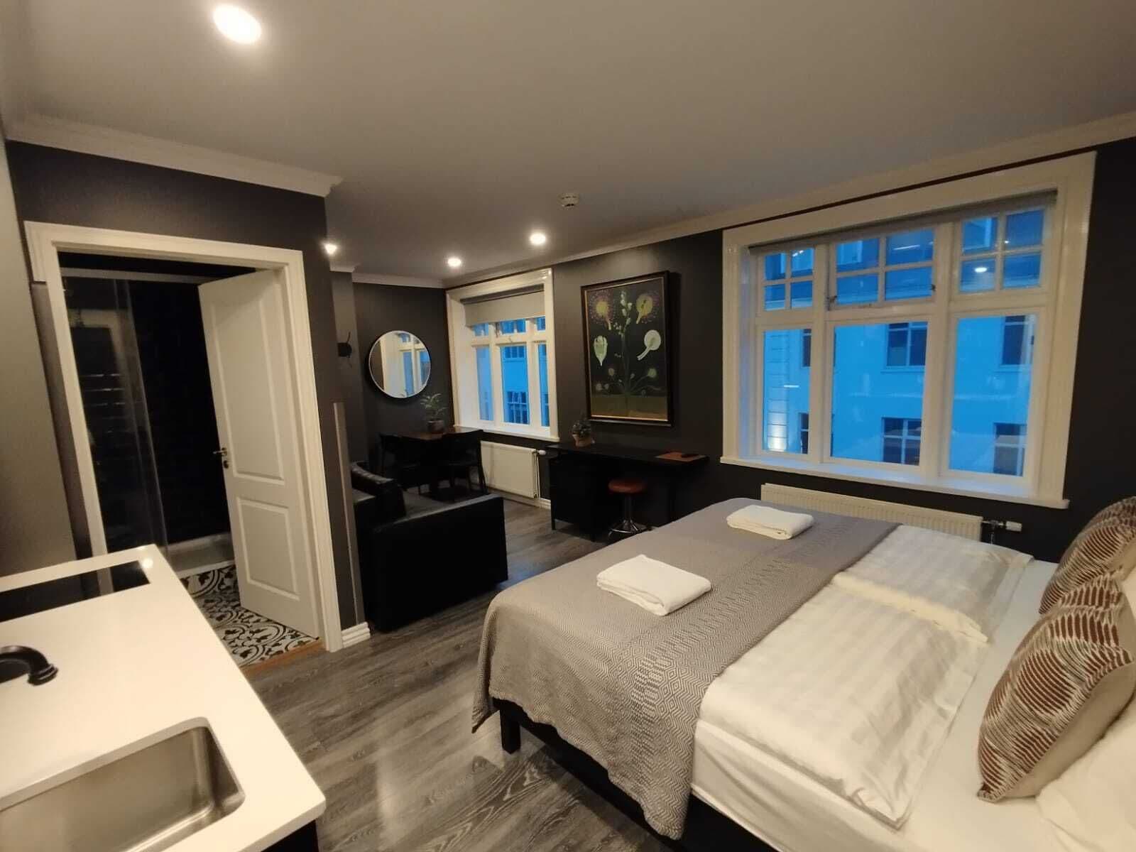 A bedroom with a bed, bedside table, and a window with a view of buildings.