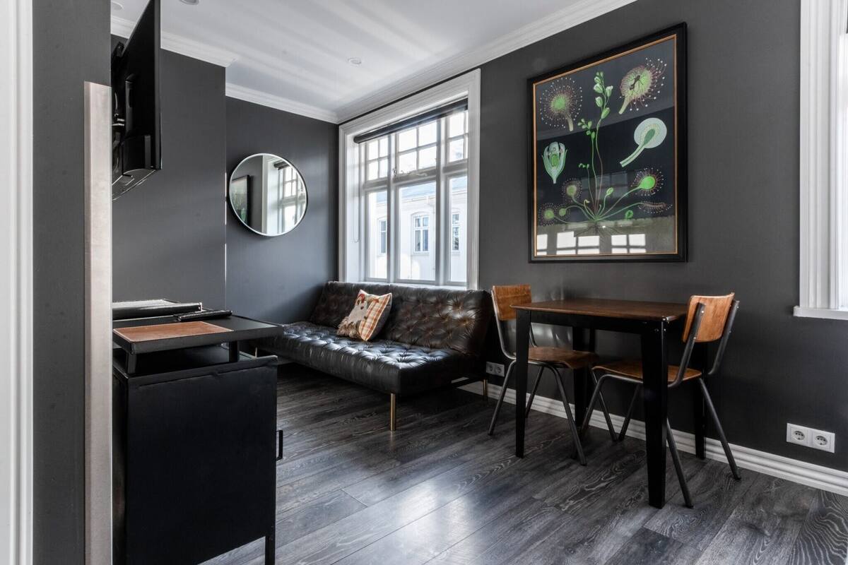 Ein modernes Zimmer mit dunklem Holzboden, einem schwarzen Ledersofa mit Kissen, einem runden Spiegel, einem Esstisch mit Stühlen und einem großen botanischen Bild im Rahmen an der Wand.