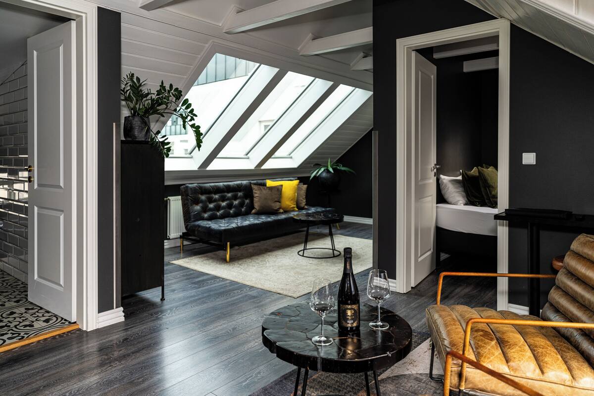 Ein modernes Wohnzimmer mit einem schwarzen Ledersofa, einem Holzsesseln und einem runden Couchtisch mit einer Flasche und Gläsern. Ein Oberlicht sorgt für natürliches Licht, und im Hintergrund ist eine Treppe zu sehen.