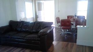 A living room with a dark brown leather sofa, a wooden floor, and a dining area with a red chair and a white table.