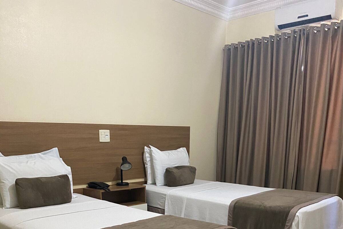 Habitación de hotel con dos camas, cada una con cubrecama marrón y blanco, cabecero de madera, mesita de noche con lámpara y una ventana grande con cortinas.