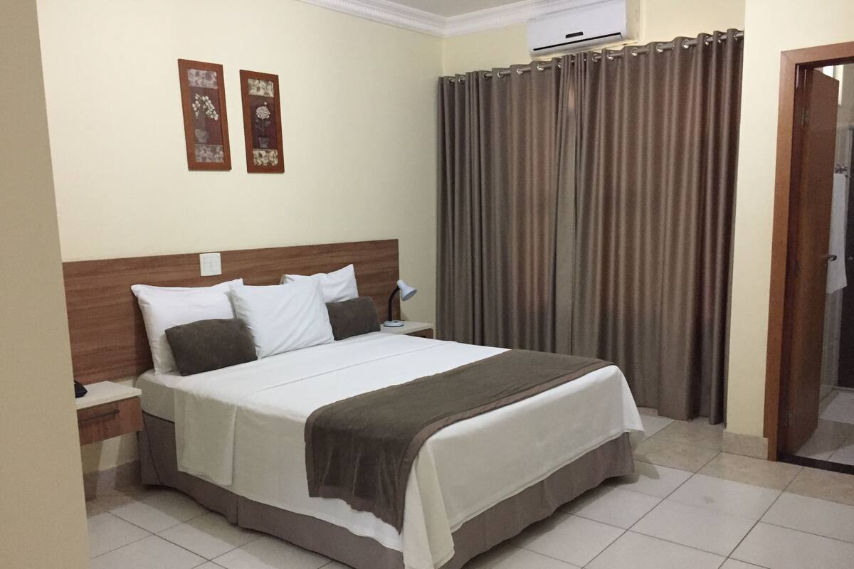 Una cama bien tendida con ropa de cama blanca y marrón, un cabecero de madera, una mesita de noche con una lámpara y una cortina en la ventana.