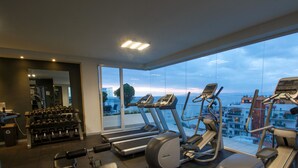 Un gimnasio con cintas de correr y mancuernas, amplias ventanas con vistas a la ciudad y un espejo que refleja los equipos de ejercicio.