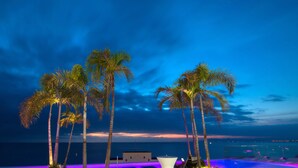 Escena de playa con palmeras y una zona de piscina al anochecer.