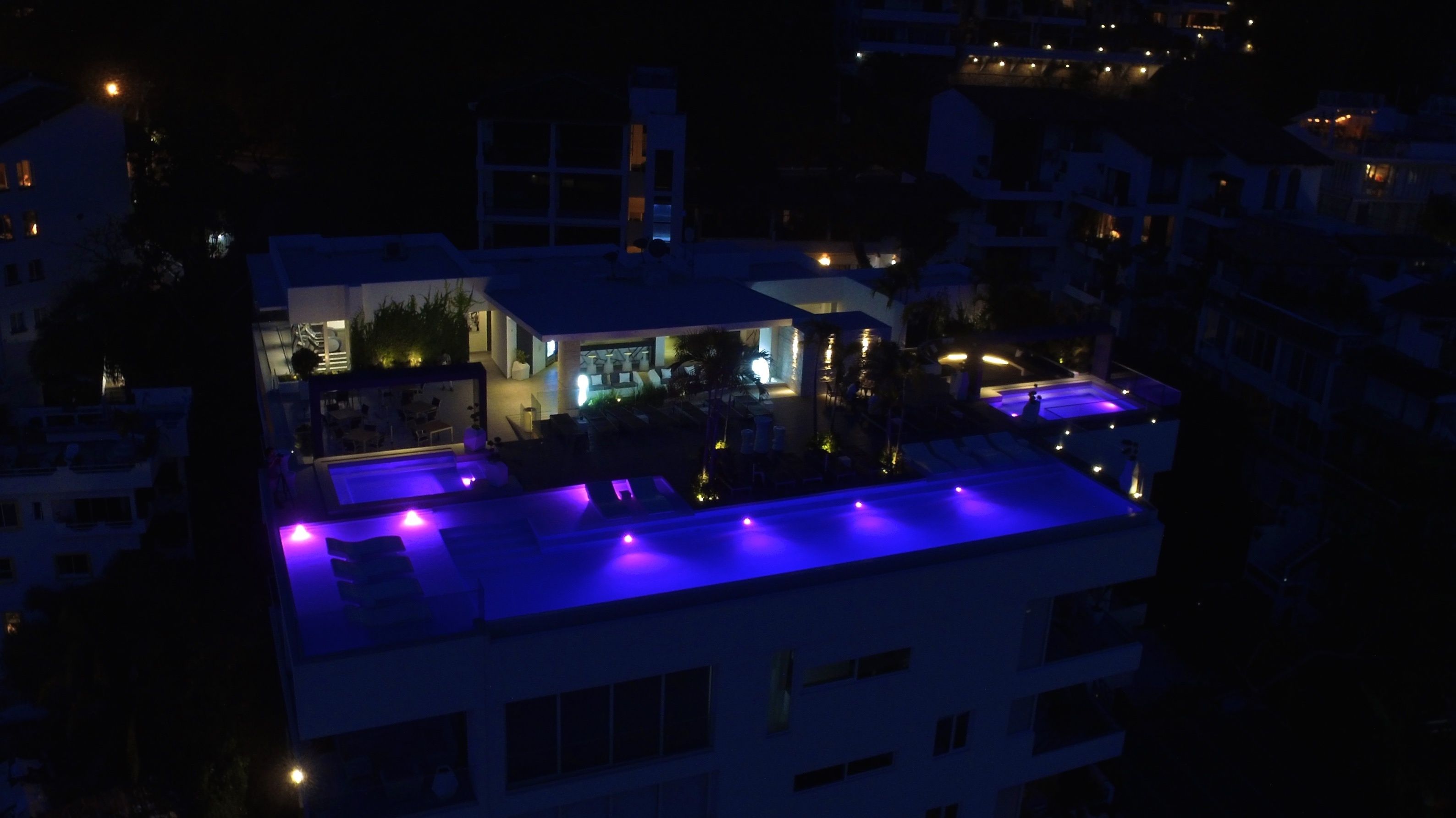 A building with a rooftop pool area illuminated by blue lights, surrounded by other buildings.