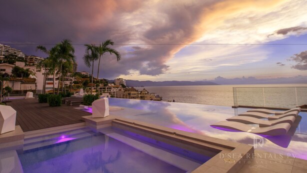Una piscina infinita en la azotea con vistas al mar y a las montañas, palmeras y una terraza de madera.