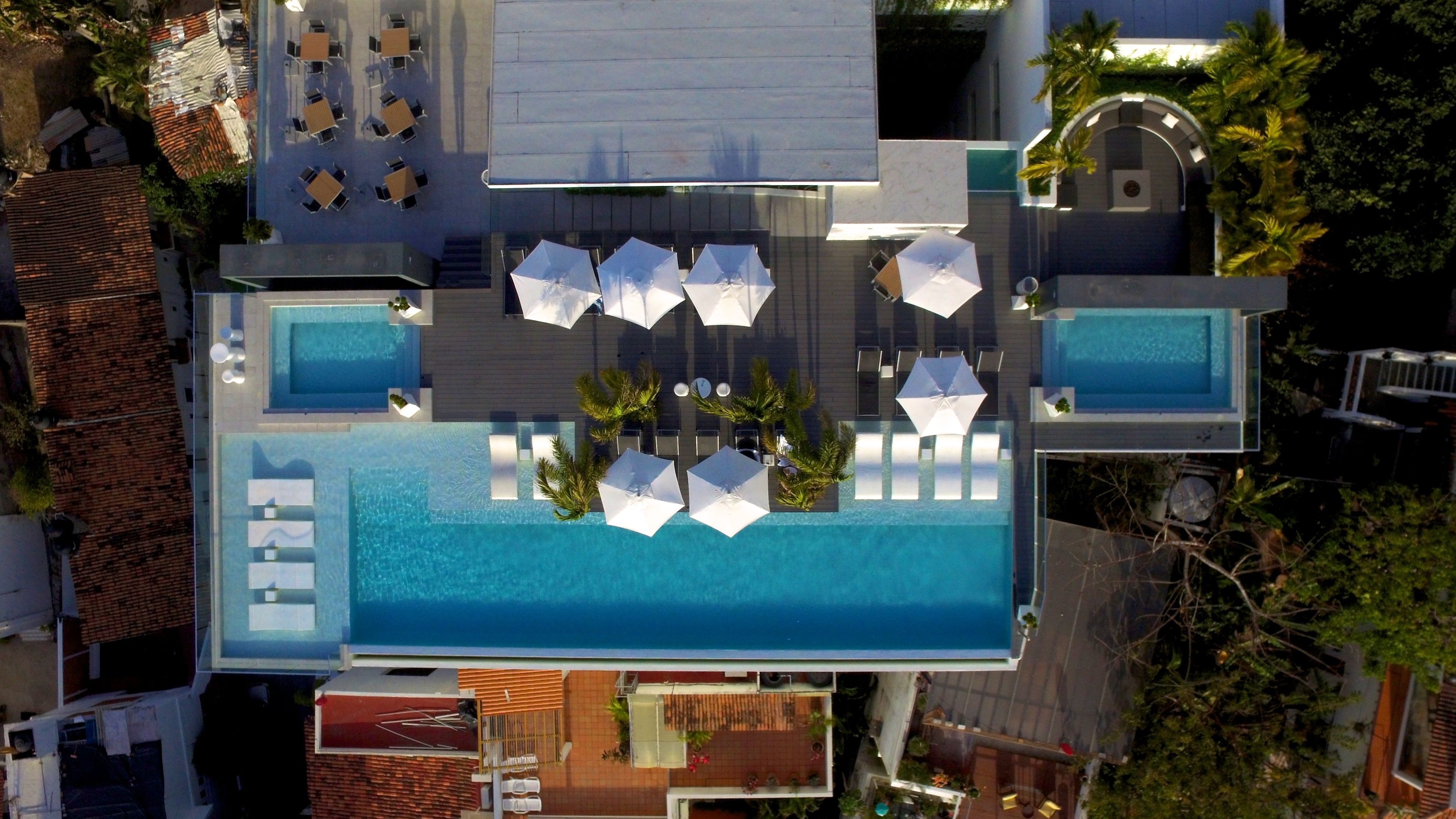 An aerial view of a hotel with a large swimming pool, multiple lounge areas with umbrellas, and a separate pool area.