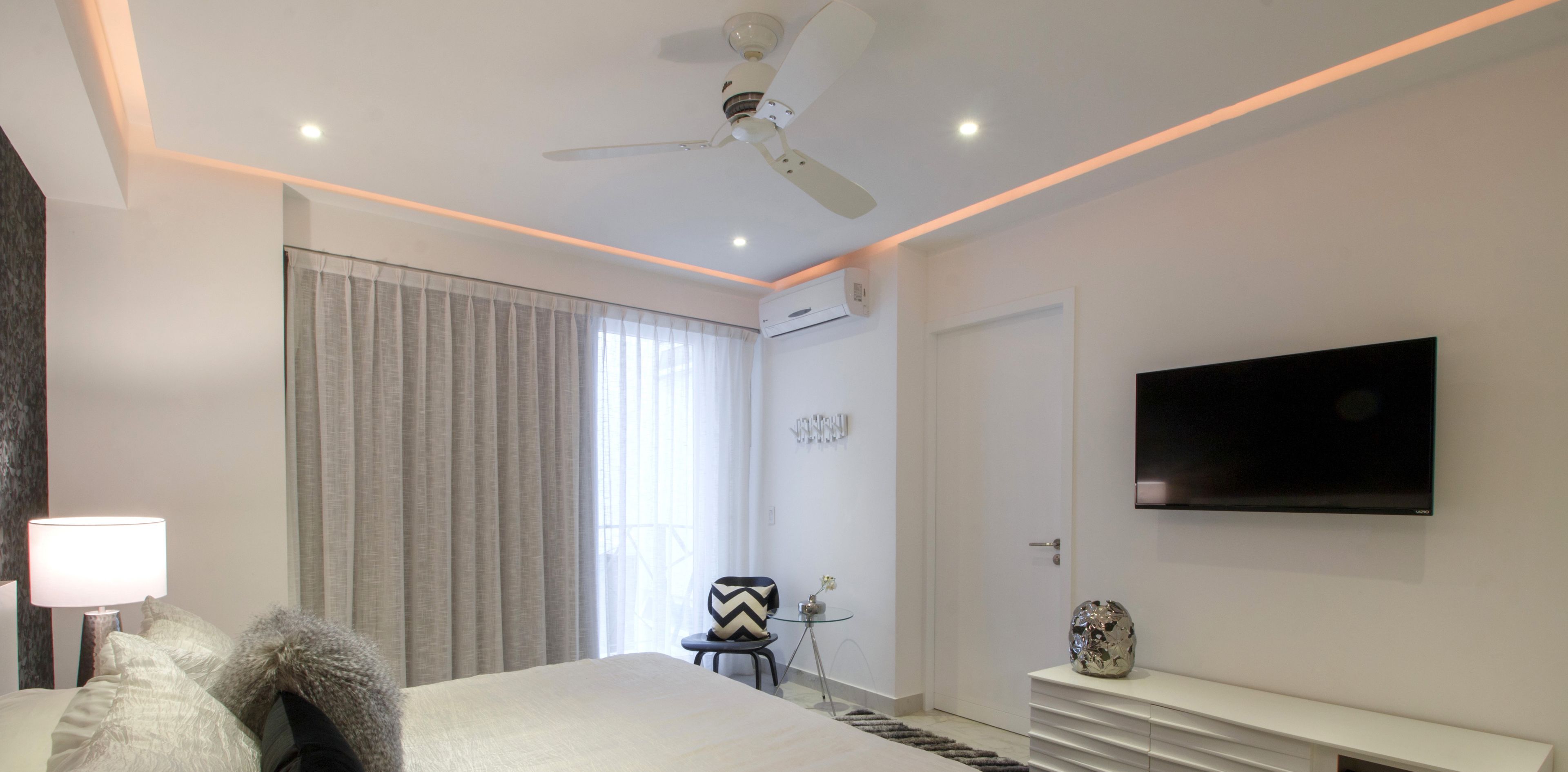 Una habitación de hotel moderna con una cama, un televisor montado en la pared, un ventilador de techo y una ventana con cortinas.