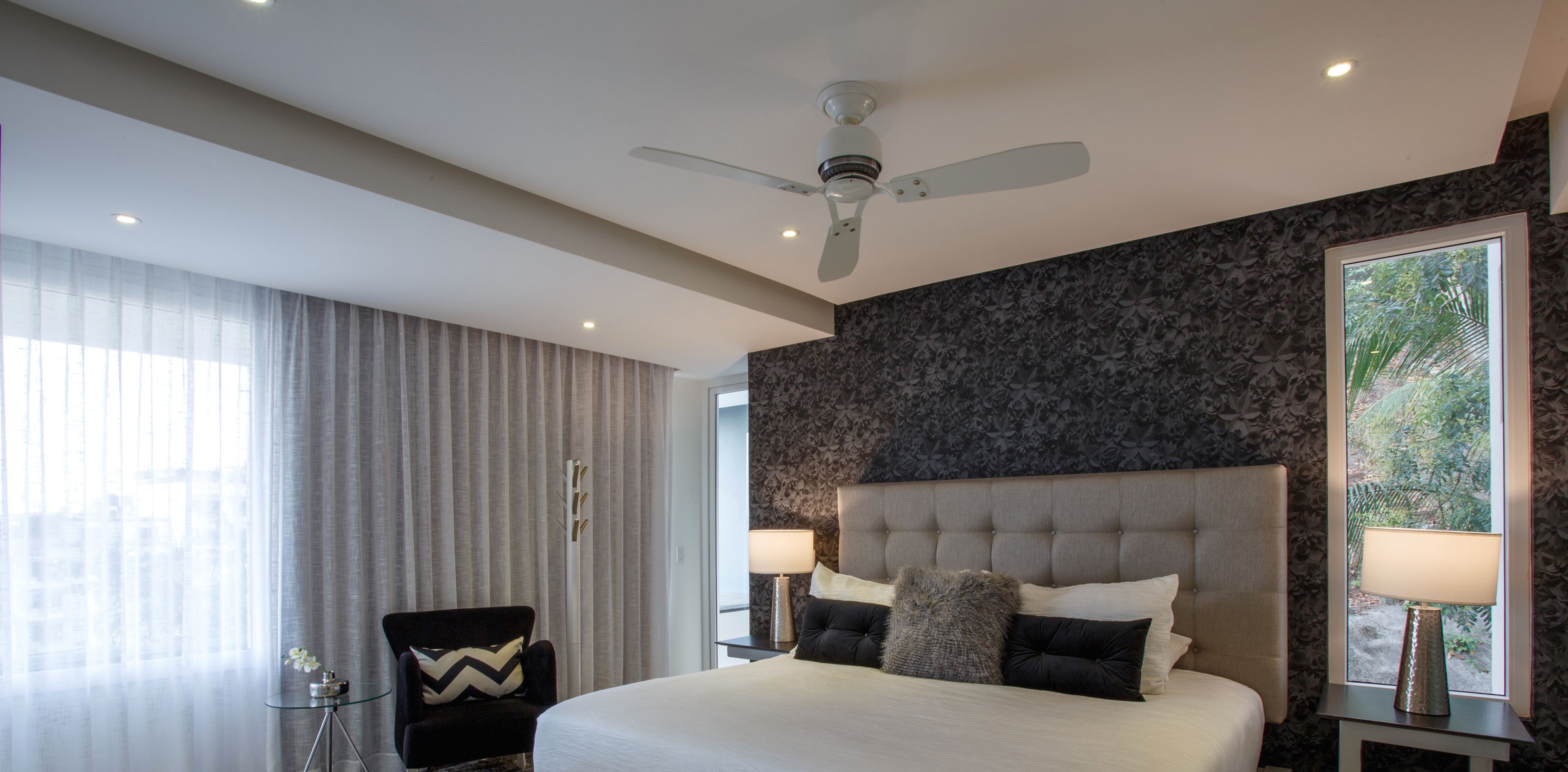 Quarto de hotel moderno com uma cama grande, um ventilador de teto, uma janela com vista e uma mesa de cabeceira com abajur.