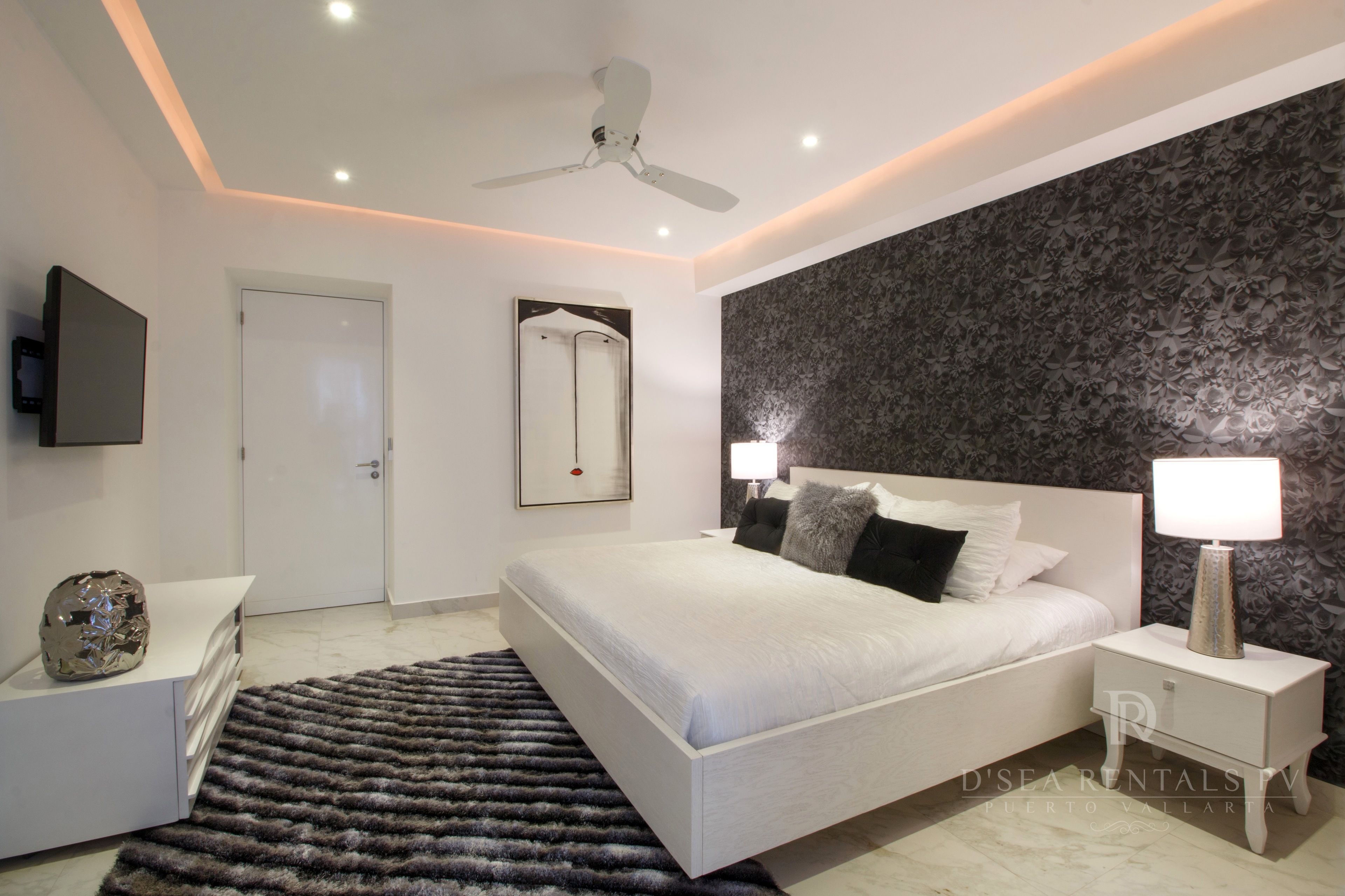 A modern bedroom with a large bed, bedside tables, a ceiling fan, and a decorative mirror.