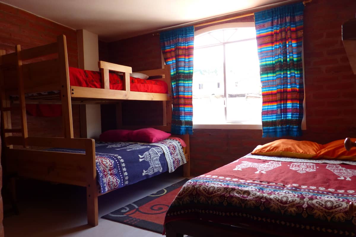 A dormitory room with two wooden bunk beds, a window with colorful curtains, and a patterned rug on the floor.