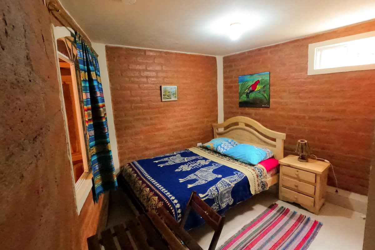 A bedroom with a brick wall, a bed with a blue bedspread, a wooden nightstand, a chair, and a colorful rug.