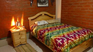 Quarto com cama de madeira, mesa de cabeceira com abajur, colcha estampada e parede de tijolos.