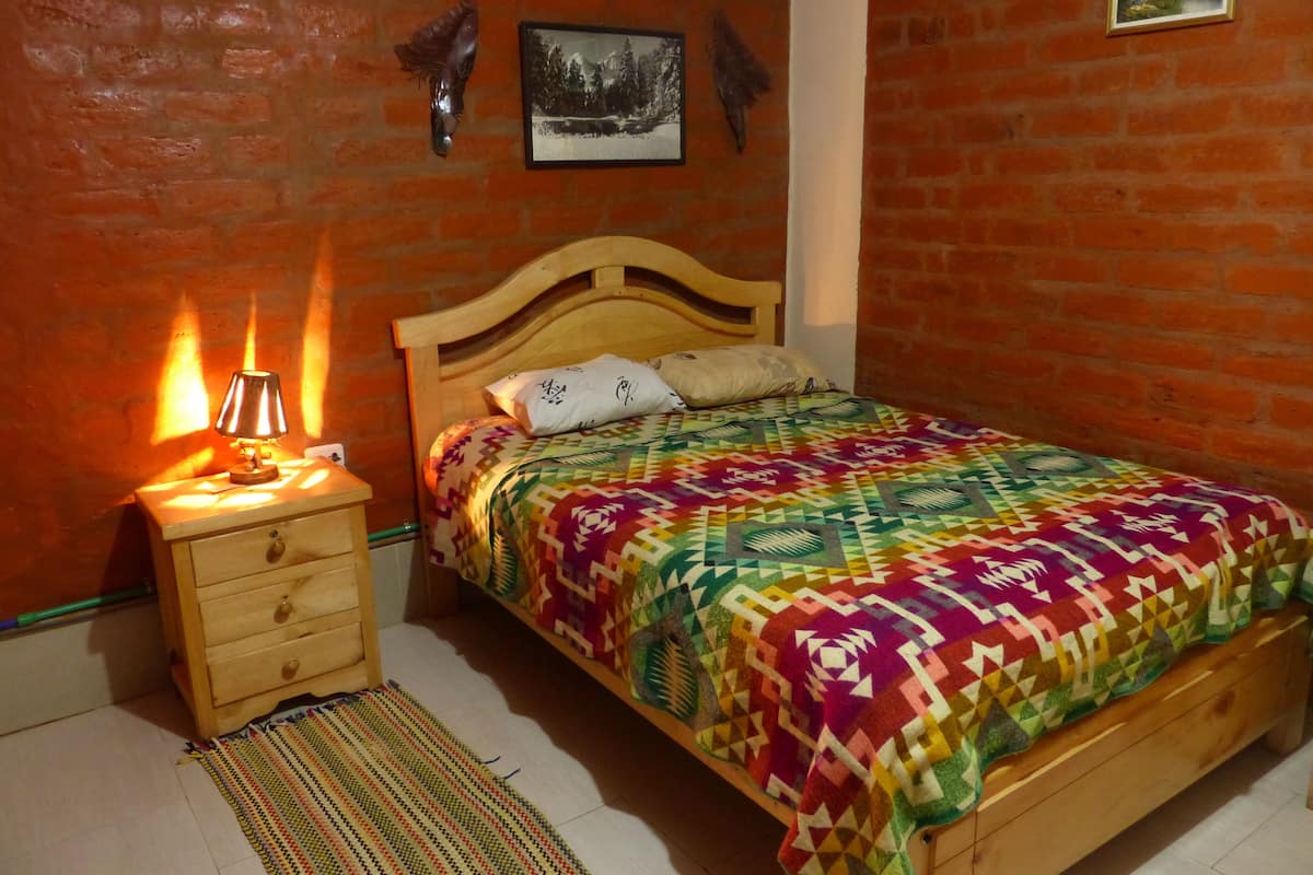 A bedroom with a wooden bed, a nightstand with a lamp, a patterned bedspread, and a brick wall.