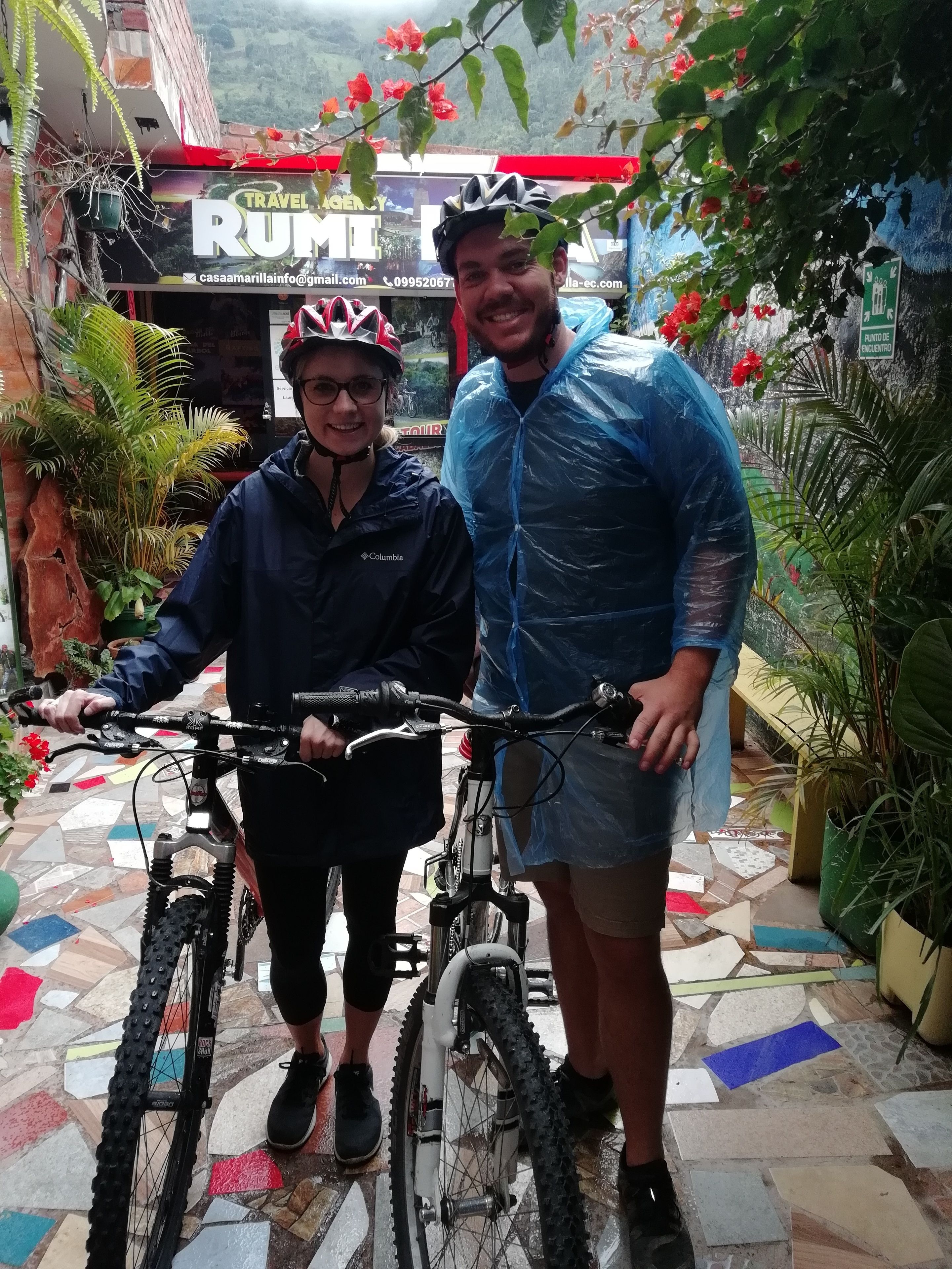 Two people standing next to bicycles in front of a building with a sign that reads "TRAVEL RUMBLE".