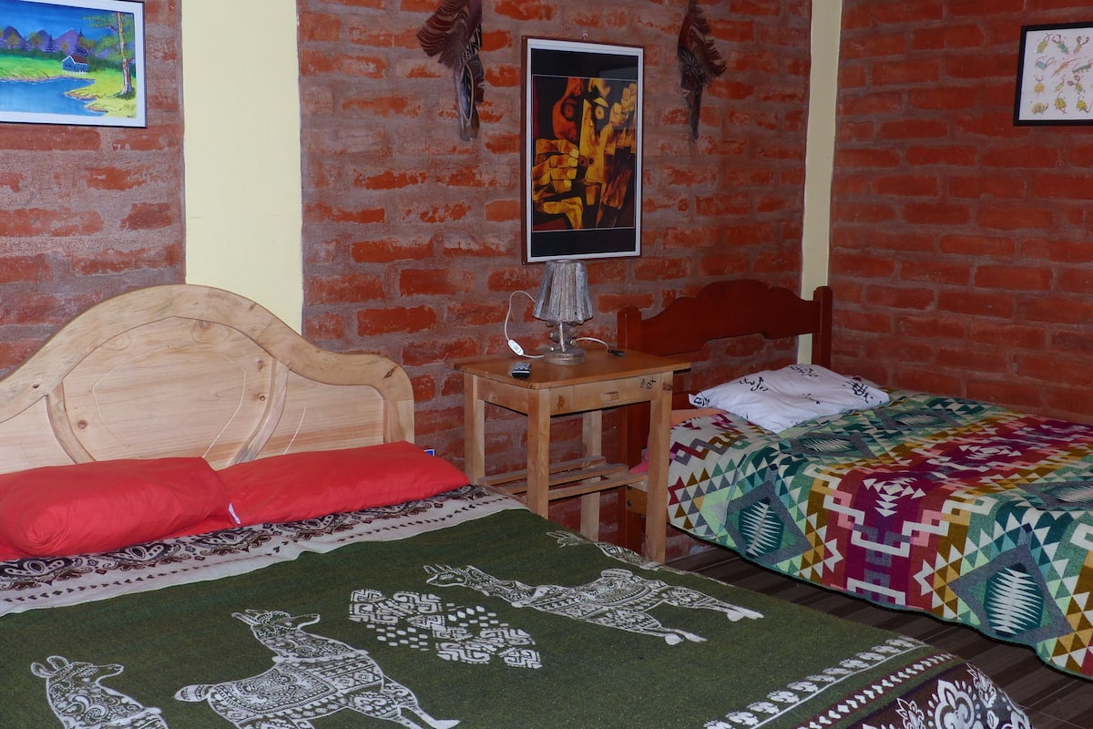 A room with a brick wall, a wooden bed with a red pillow, a colorful quilt, and a framed picture on the wall.
