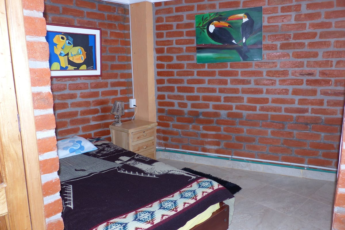 A room with a brick wall, a bed with a patterned blanket, a wooden nightstand, and two framed artworks.