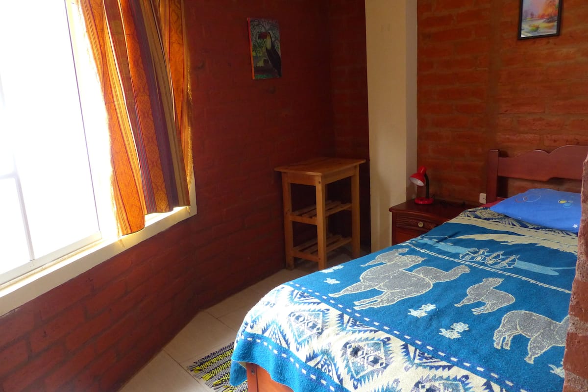 A room with a bed, a small wooden table, a red lamp, and a window with curtains.