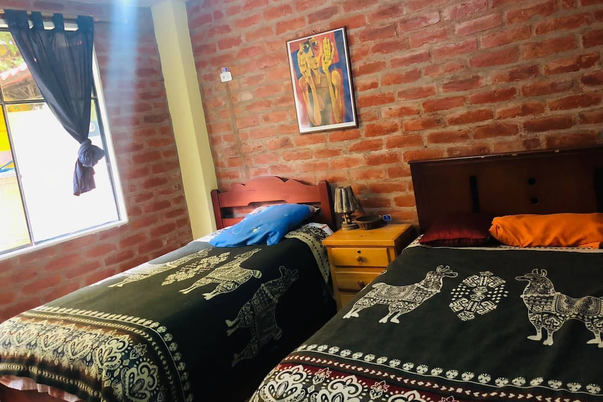 A room with a brick wall, two beds with patterned bedspreads, a nightstand with a lamp, and a window with curtains.