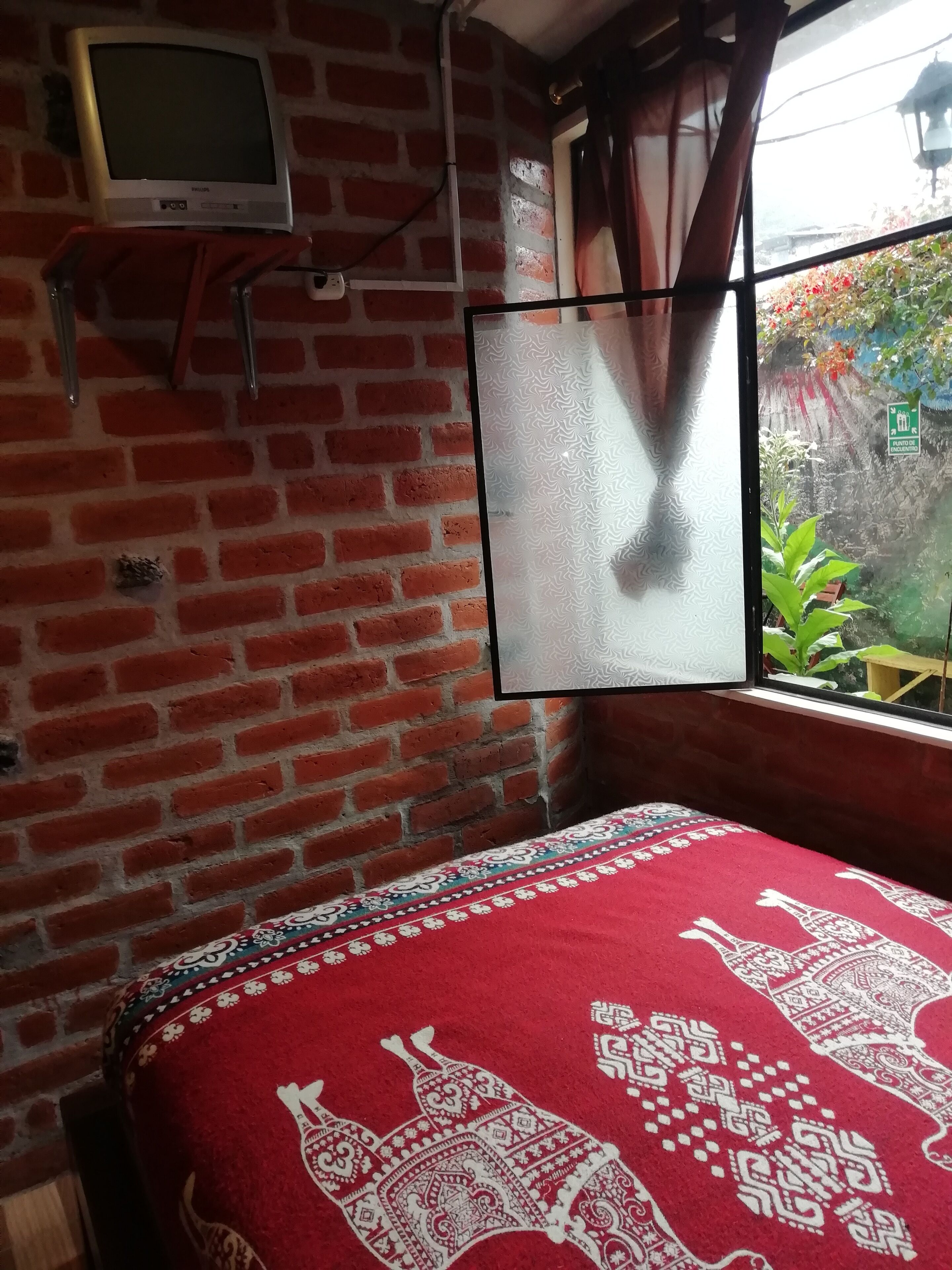 A room with a brick wall, a bed with a red patterned bedspread, a television on a shelf, and a window with a view of greenery.