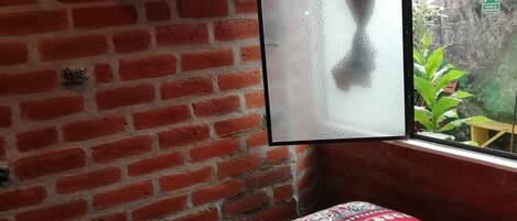 A room with a brick wall, a bed with a red patterned bedspread, a television on a shelf, and a window with a view of greenery.