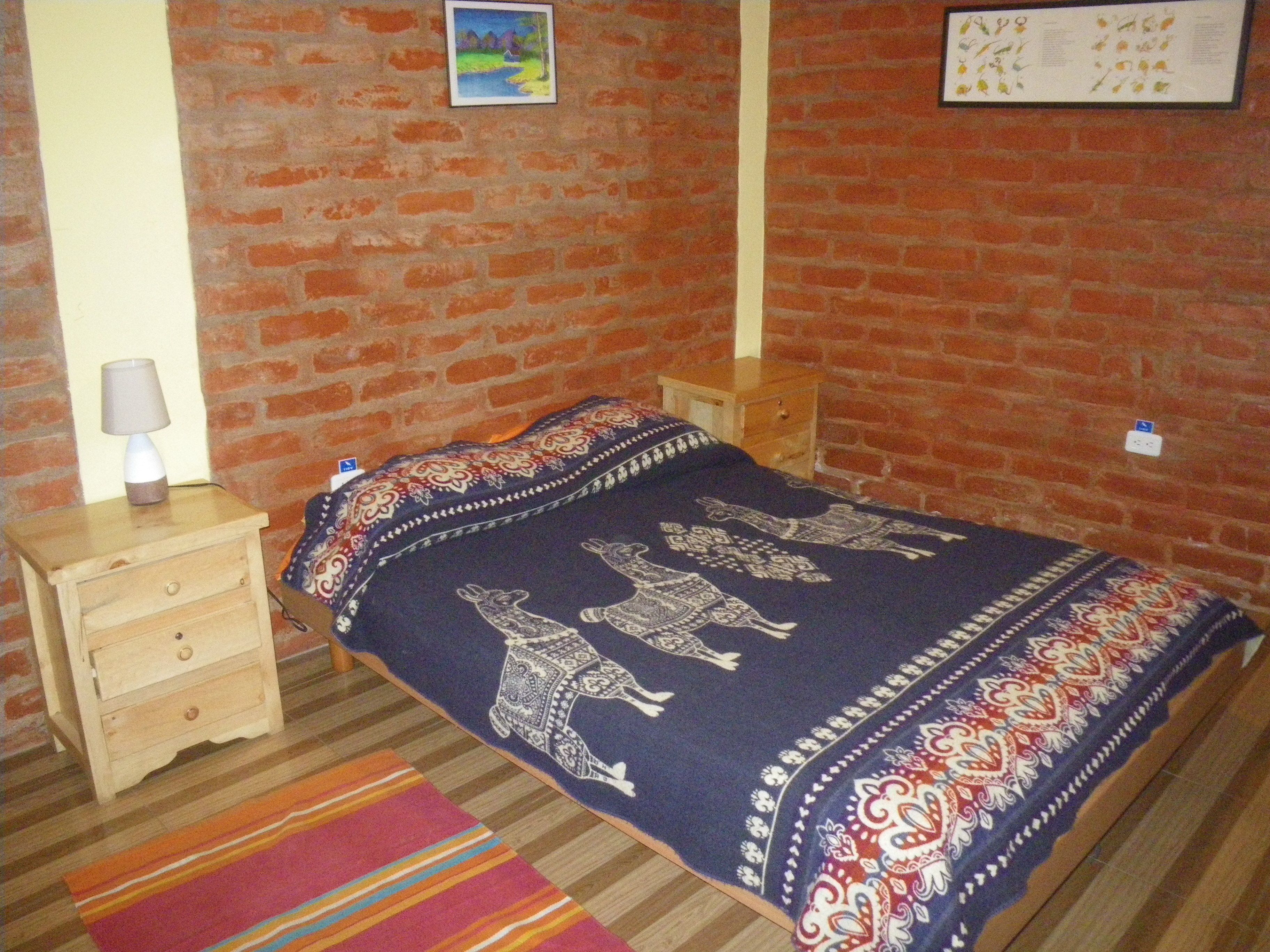 A room with a brick wall, a wooden nightstand with a lamp, a bed with patterned bedding, and a colorful rug.