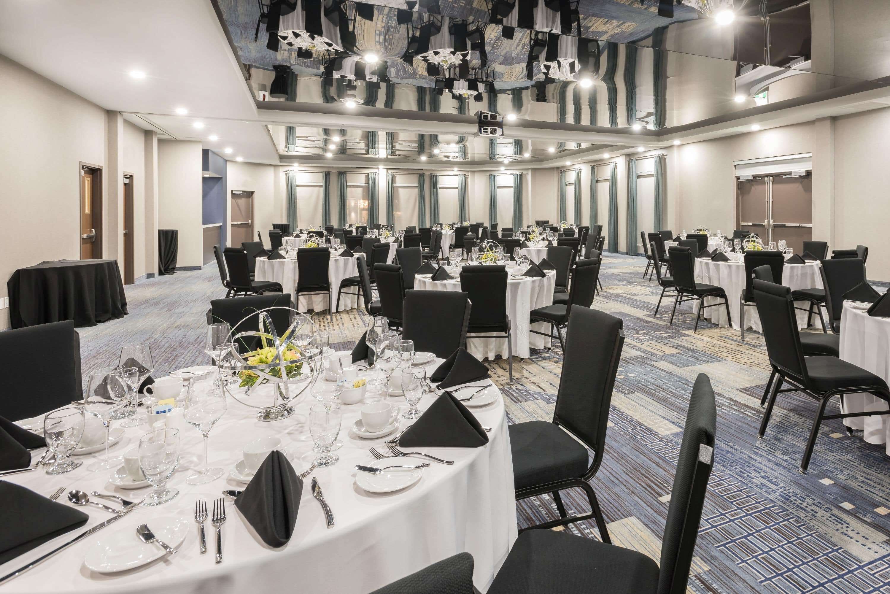 Un amplio salón de conferencias con varias mesas redondas dispuestas para comidas, equipado con manteles blancos, sillas negras y cubertería.