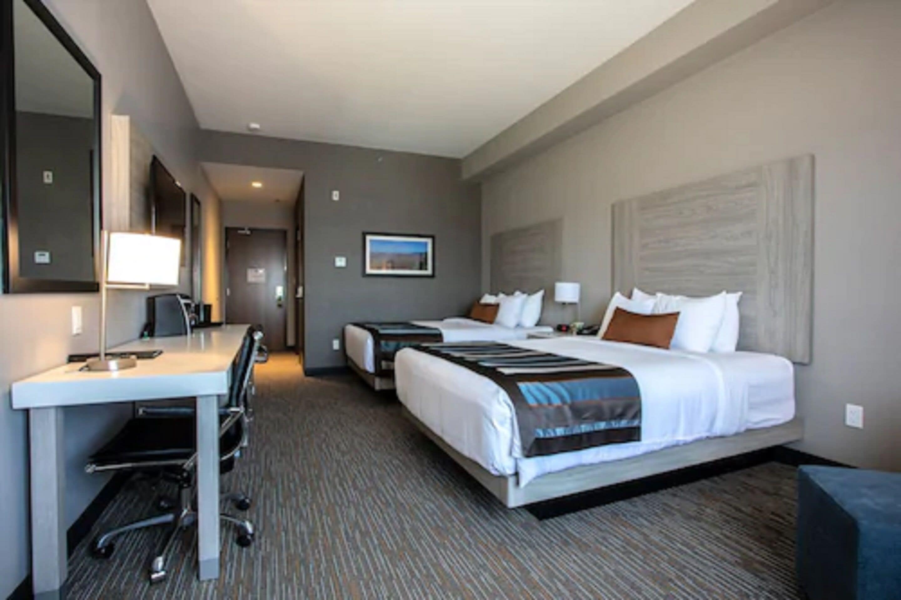 A hotel room with a desk, two beds, a large mirror, and a framed picture on the wall.