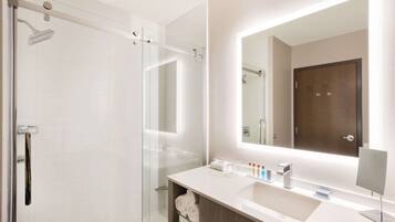 Un baño moderno con ducha envuelta en vidrio, un espejo grande y un lavabo blanco con tocador de madera.