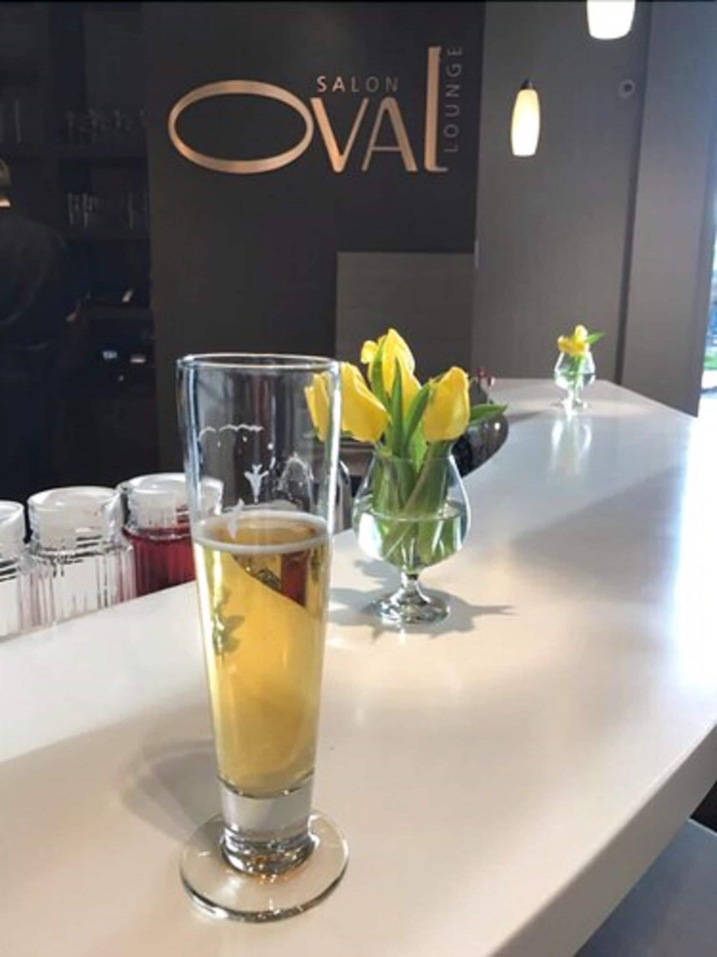 A bar area with a tall glass of a yellow beverage, a small vase with yellow flowers, and a sign reading "SALON OVAL LOUNGE".