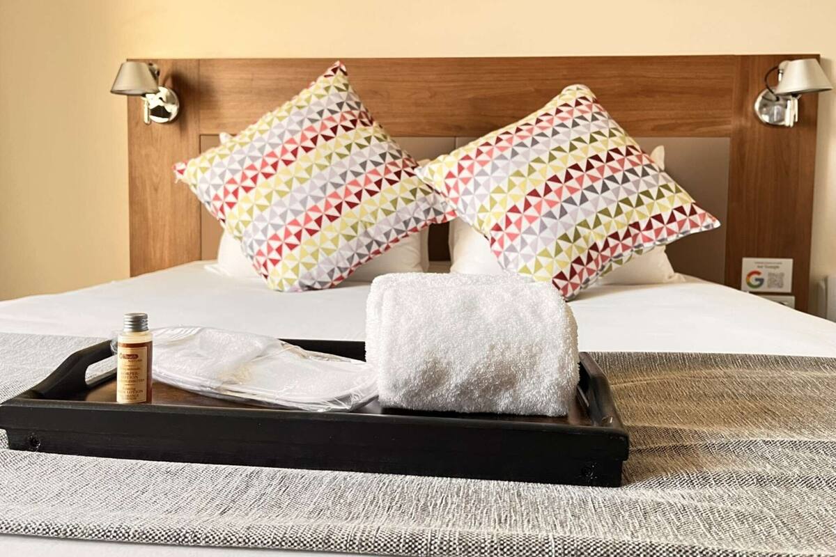 Un lit bien fait, agrémenté d’oreillers à motifs, une tablette avec des articles de toilette et une tête de lit ornée de lampes fixées au mur.