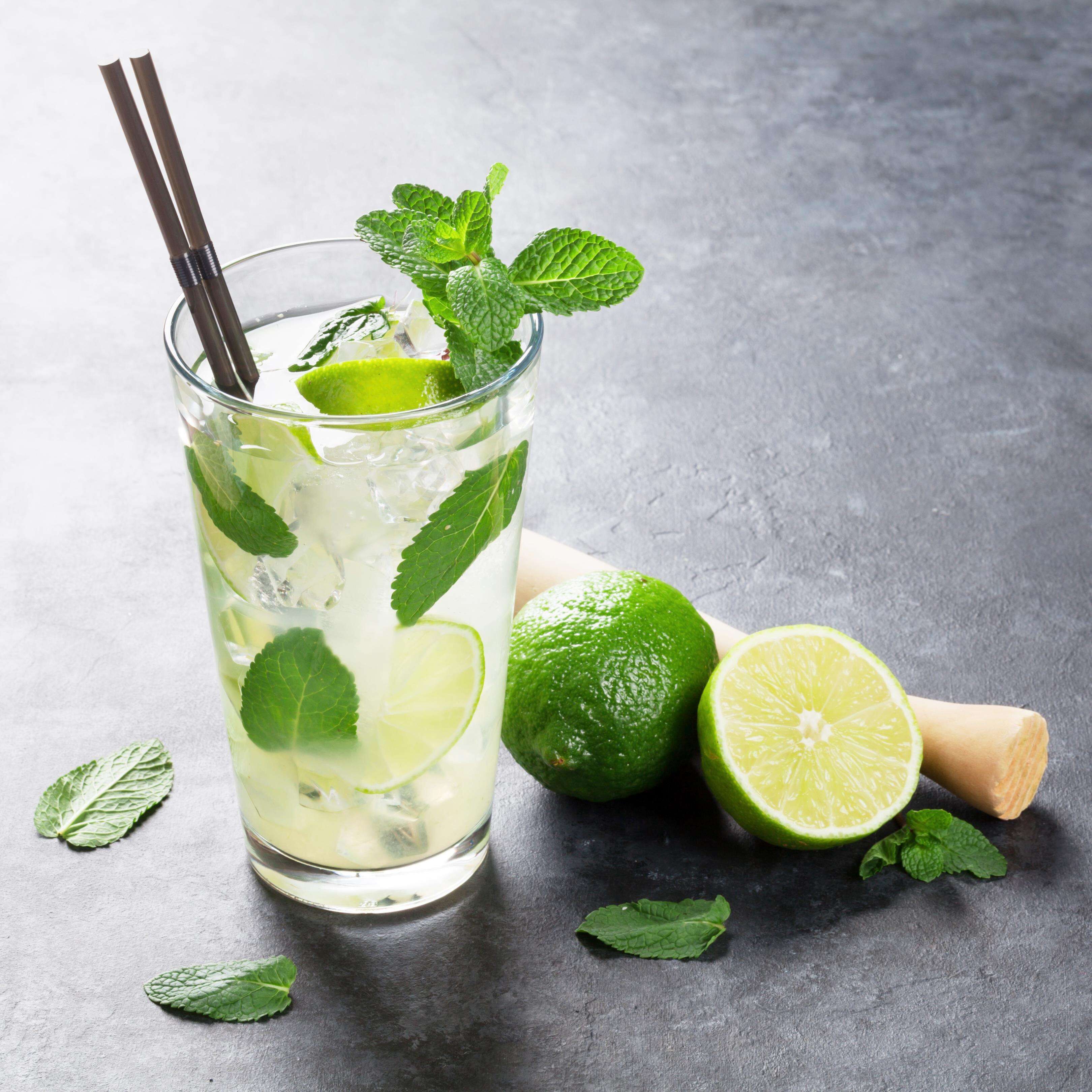 Un vaso de bebida a base de lima con hojas de menta y cubitos de hielo, acompañado de limas enteras y a la mitad sobre una superficie con textura.