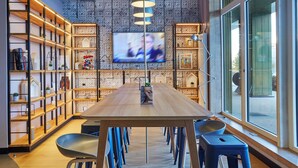 A modern meeting room with a long table, chairs, and built-in shelving. A television is mounted on the wall, and there are large windows providing natural light. - Campanile PRIME - Paris Saclay (Gif-sur-Yvette)