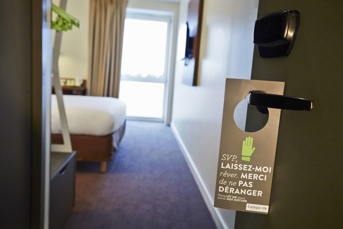 Une chambre d’hôtel avec un lit, une table de chevet et une fenêtre avec des rideaux. Une pancarte sur la porte indique : « SWP, LAISSEZ-MOI rêver. MERCI de ne PAS déranger » et « Veuillez, LAISSEZ-MOI rêver et NE PAS déranger ».