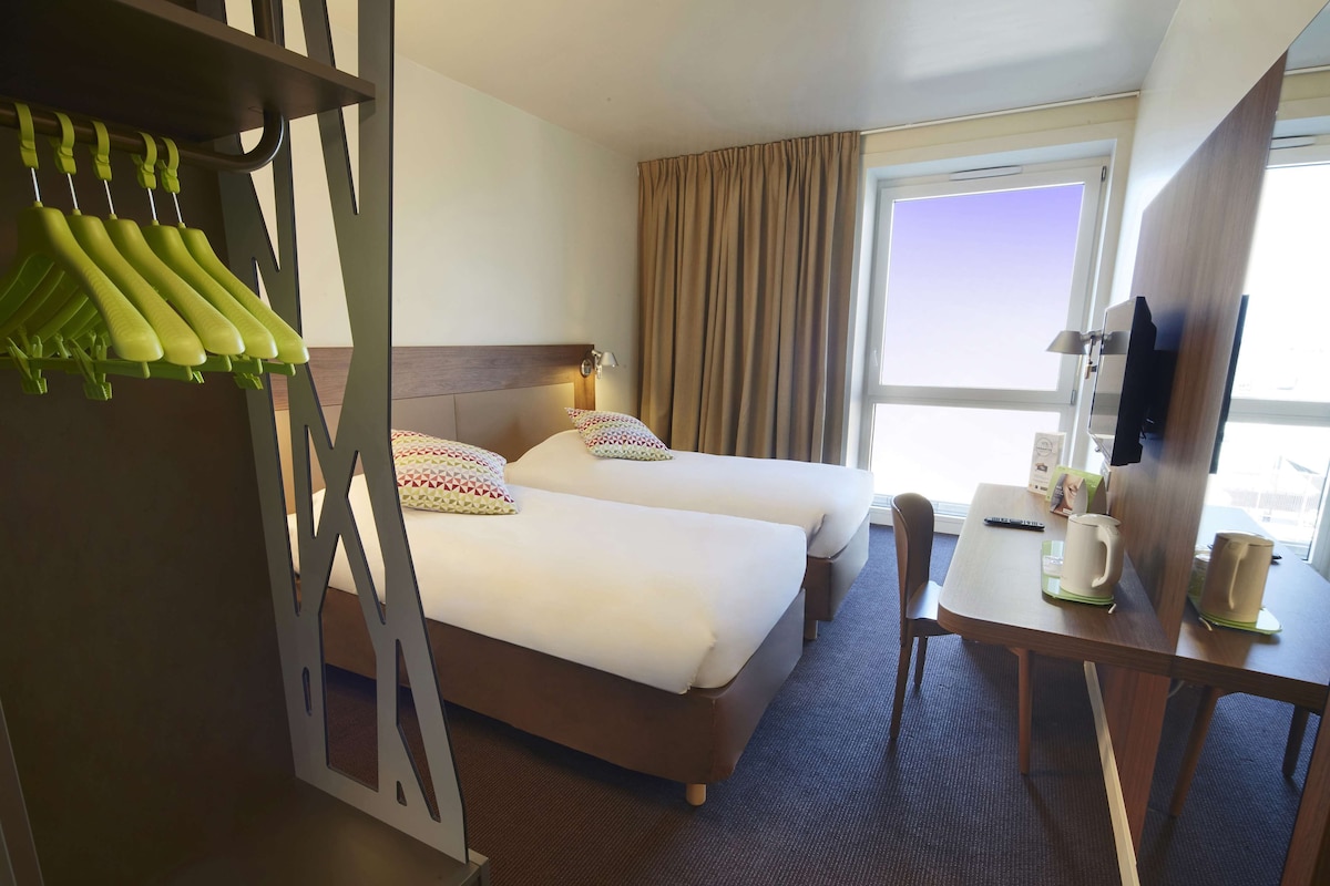 Une chambre d’hôtel avec deux lits, un bureau avec une chaise, une télévision et une fenêtre avec des rideaux.