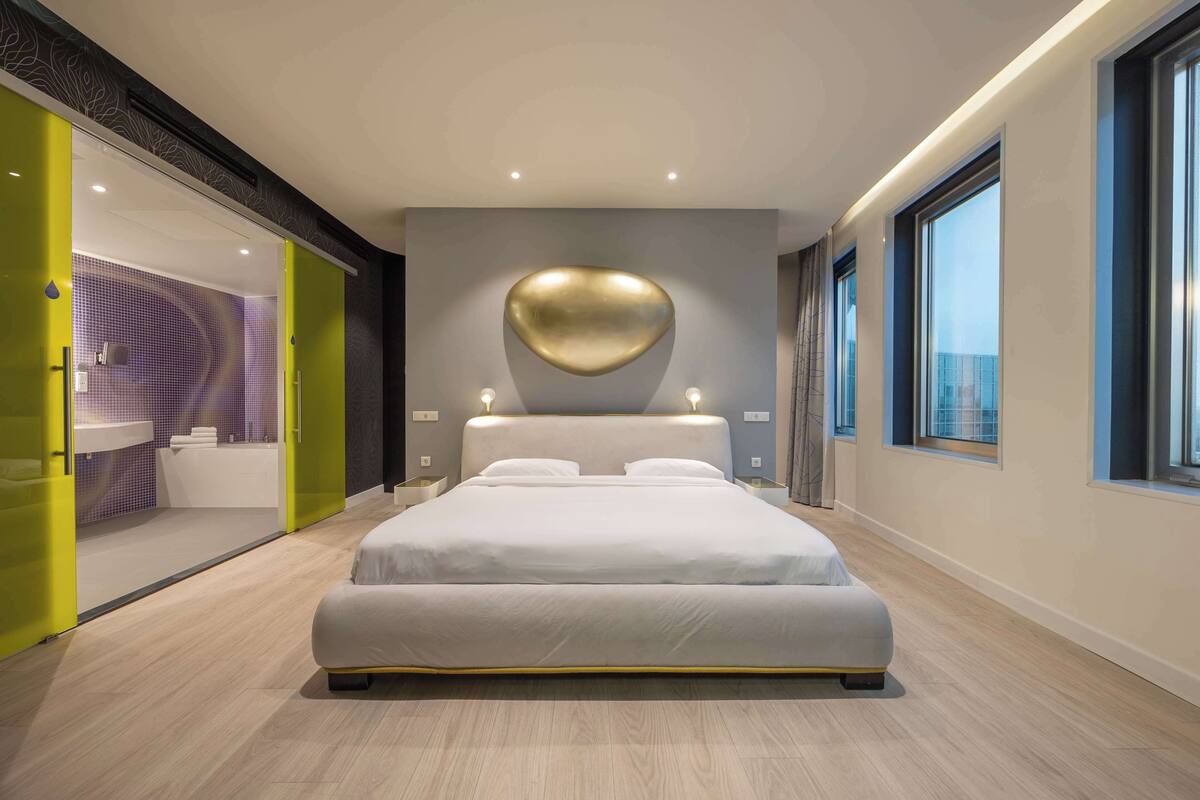 A modern bedroom with a large bed, a heart-shaped wall decoration, and a view of the city through large windows.