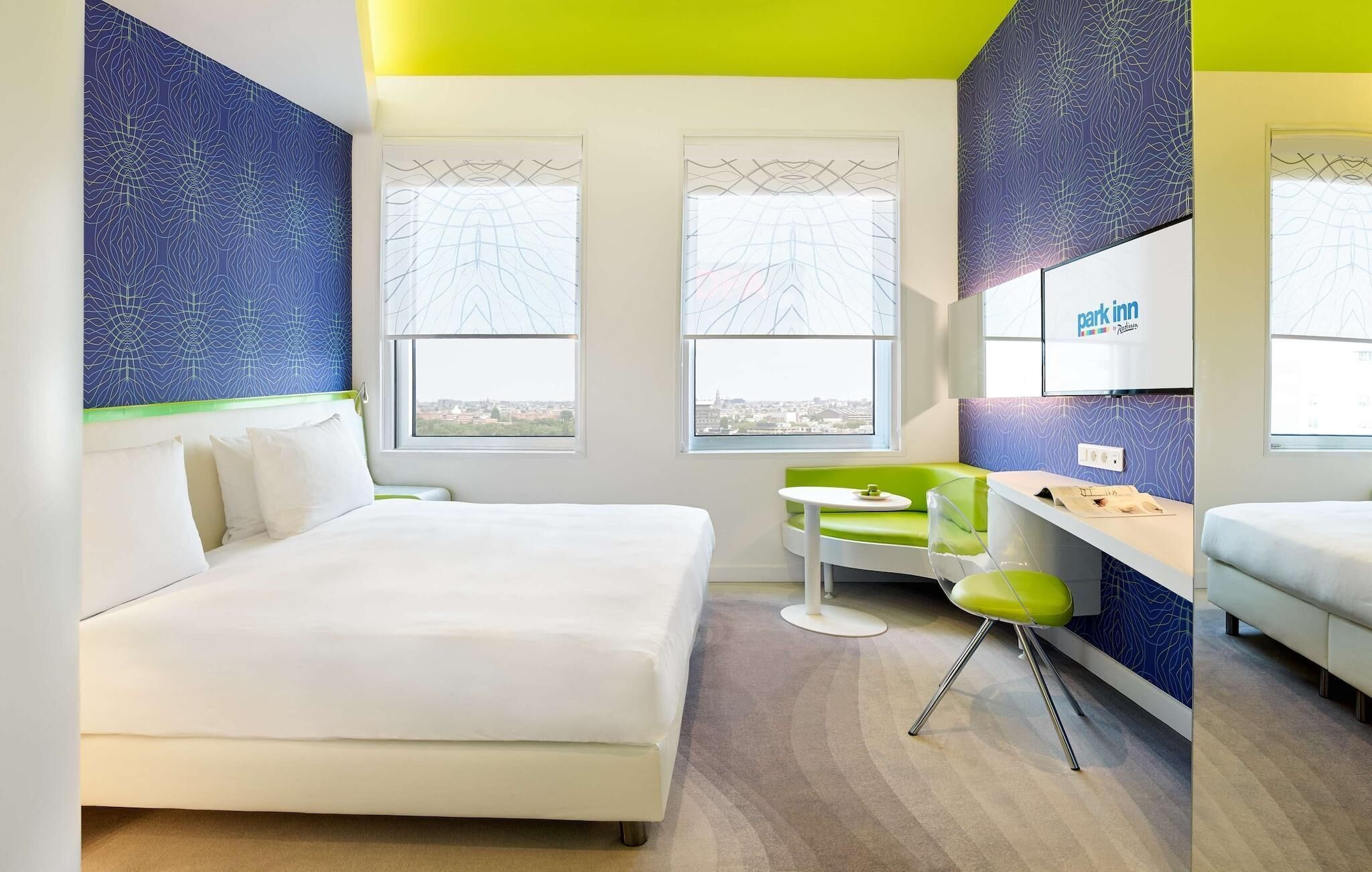 Une chambre d’hôtel moderne avec un grand lit, une petite table, un fauteuil vert et une vue sur la ville.