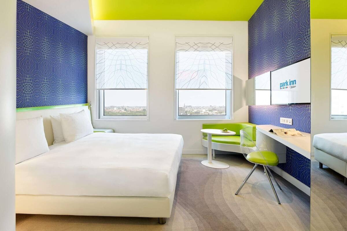 A modern hotel room with a large bed, a small table, a green chair, and a city view.