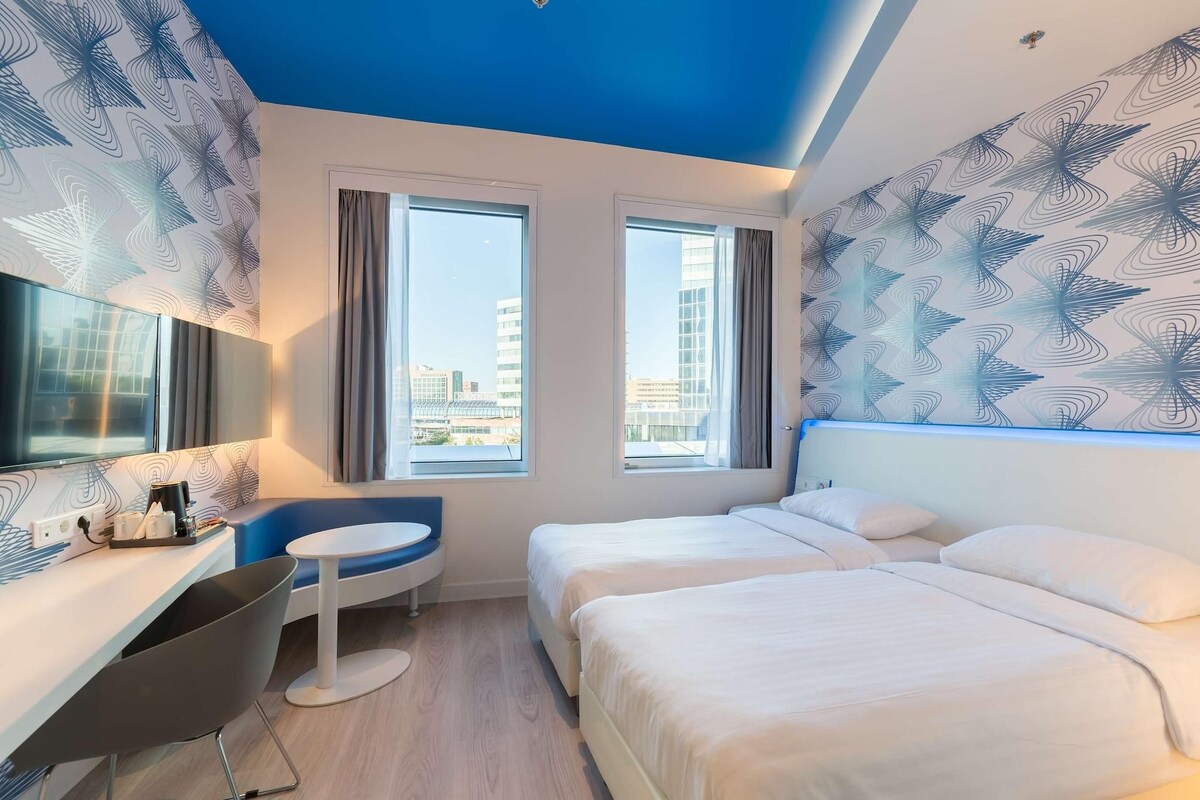A hotel room with two beds, a desk, a chair, and large windows with a city view.