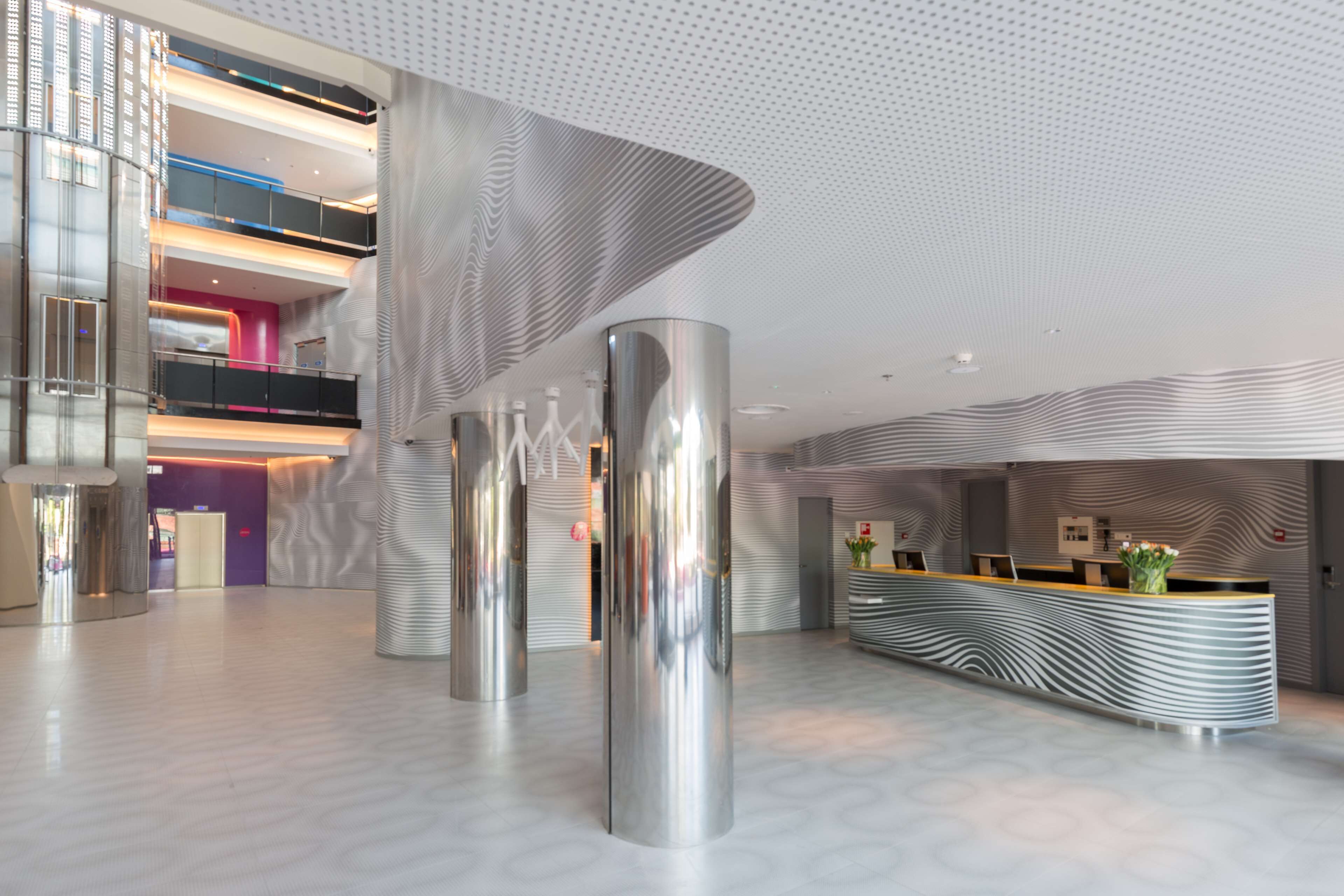 Eine moderne Hotelhalle mit einer Rezeption, metallenen Säulen und einer gemusterten Decke.