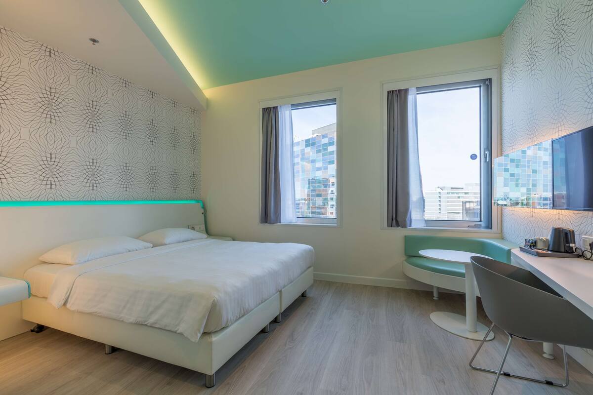 A modern hotel room with a bed, a desk, and a window with a view of the city.