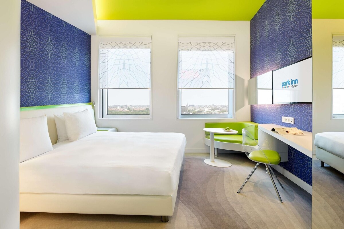 Una habitación de hotel moderna con una cama grande, una pequeña mesa, una silla verde y vistas a la ciudad.