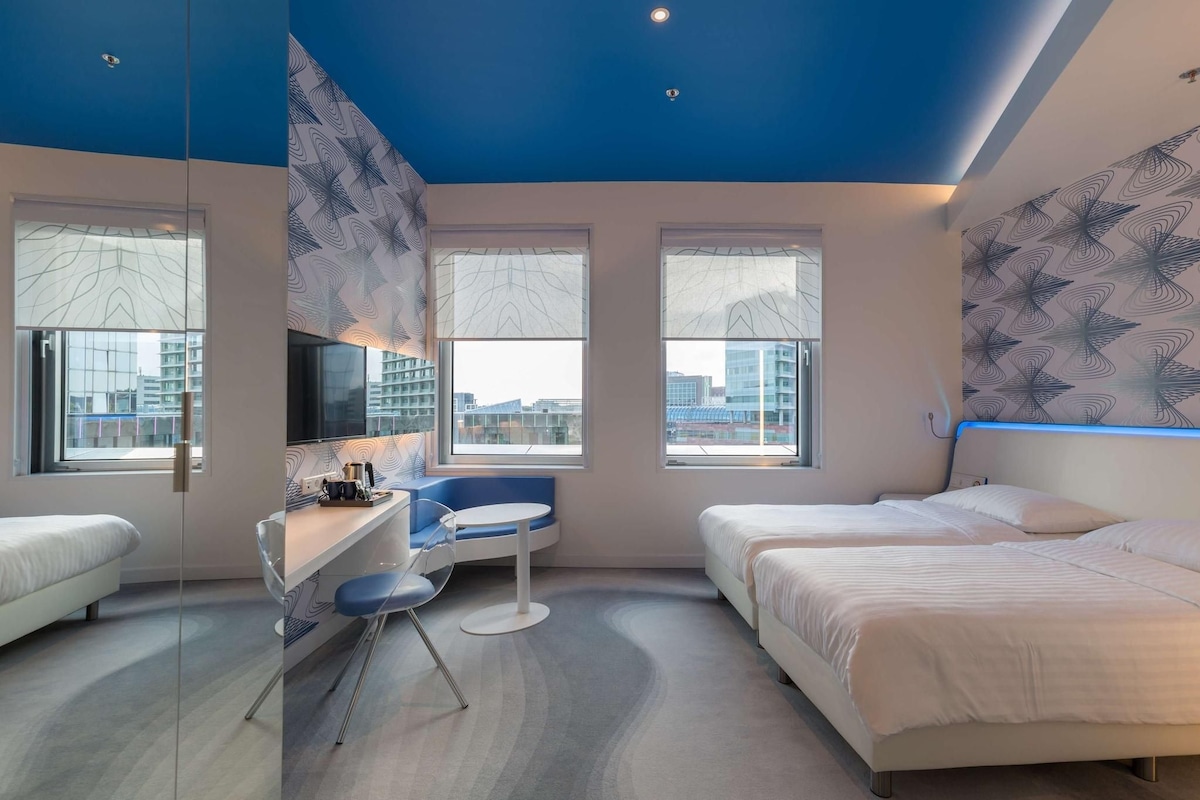 A modern hotel room with a bed, a desk, a chair, and a small table. There are large windows with a view of the city.