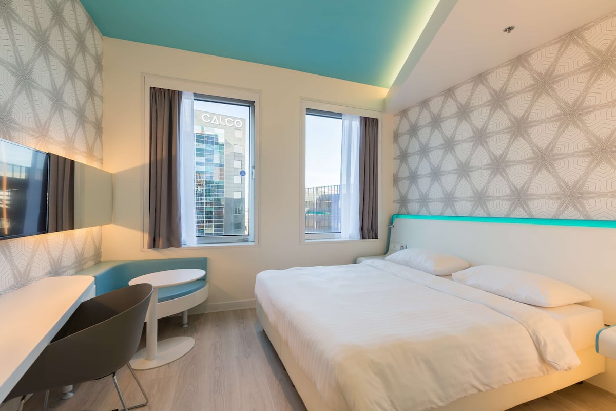 A modern hotel room with a bed, a desk, and a chair. There are two windows with curtains, and a patterned wall.