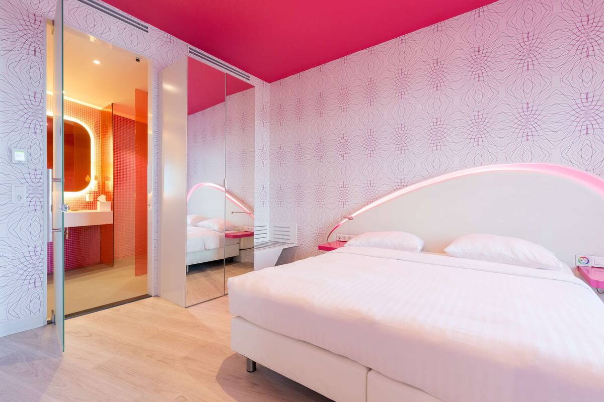 A modern bedroom with a large bed, a glass door leading to a bathroom, and a pink ceiling.