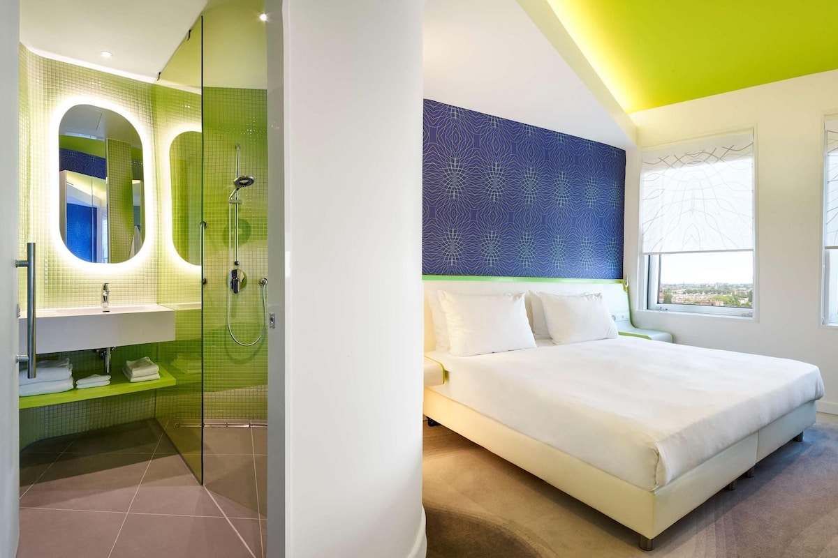 A modern hotel room with a glass-enclosed shower, a large bed, and a city view.