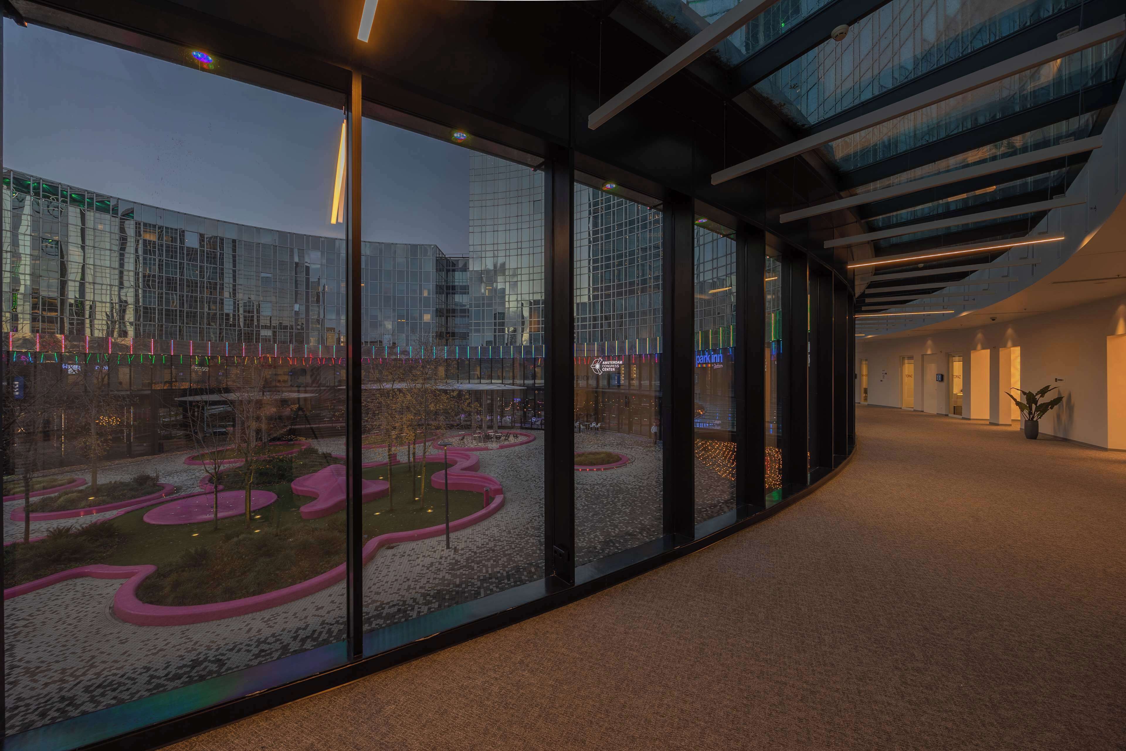 Un pasillo moderno de hotel con ventanas amplias, techo curvo y vistas a un jardín exterior.