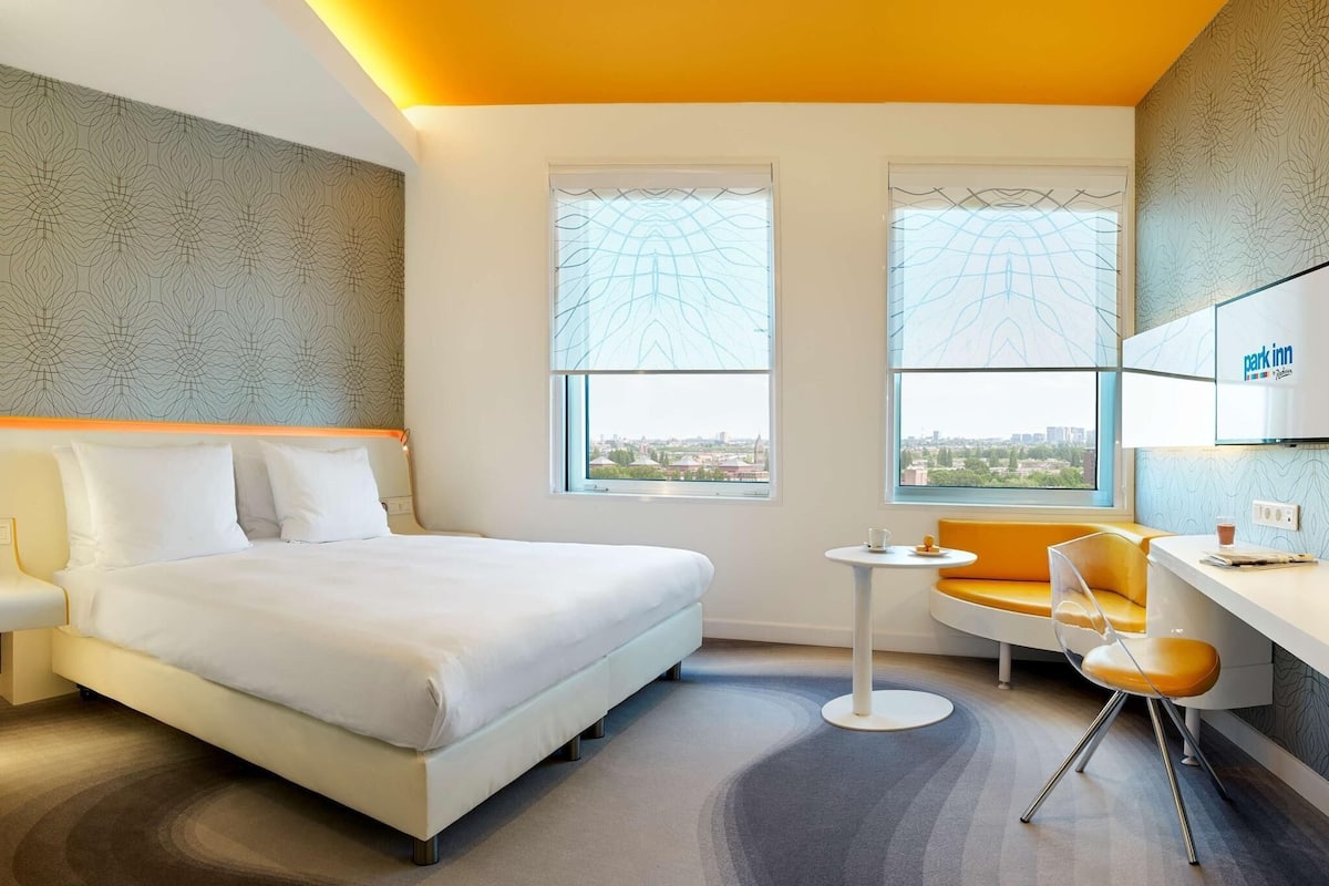 A hotel room with a bed, a desk, a chair, two windows, and a view of the city.