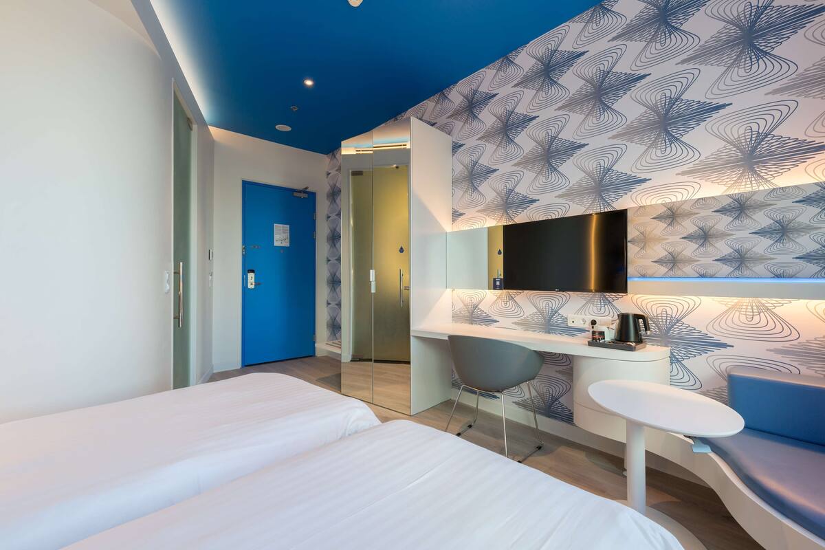 A modern hotel room with a large bed, a desk with a chair, a small table, a blue door, and a patterned wall.